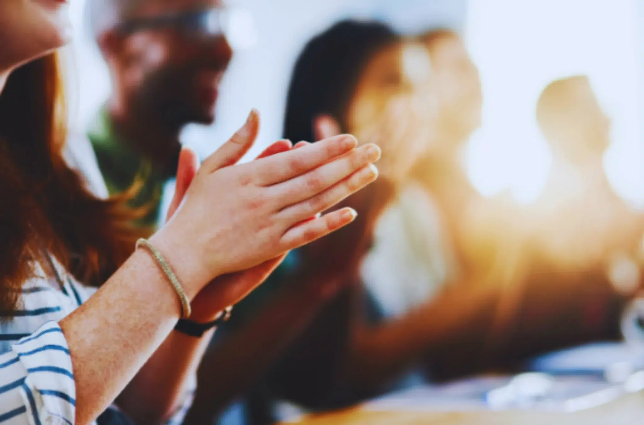 lose-up view of diverse hands clapping in a group, with other people blurred in the background, suggesting an audience or team meeting.