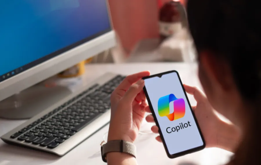 A person holding a smartphone displaying the Microsoft Copilot app logo and name on the screen, with a desktop computer and keyboard blurred in the background.