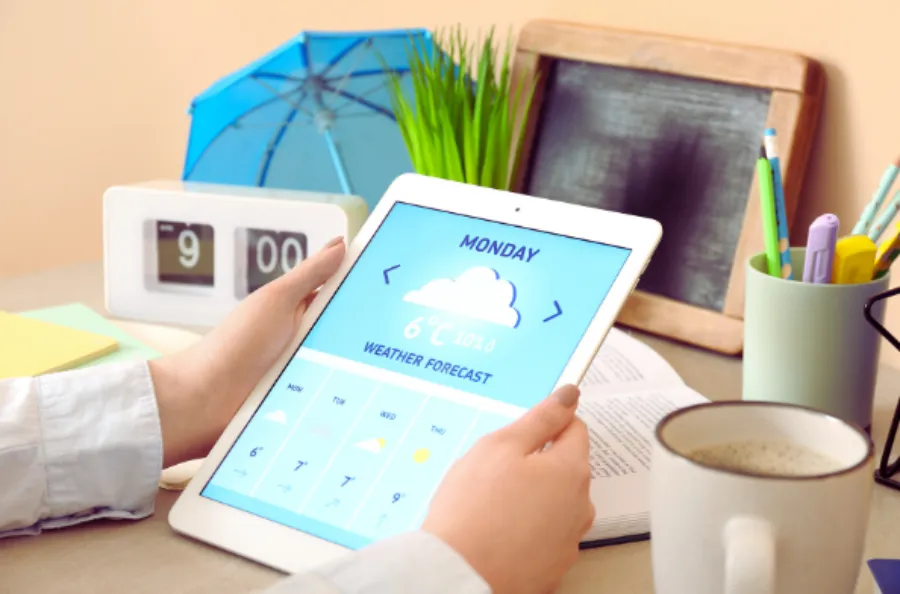 Alt Text: A person holding a tablet that displays a weather forecast app, with an alarm clock, umbrella, and coffee mug on a desk in the background.