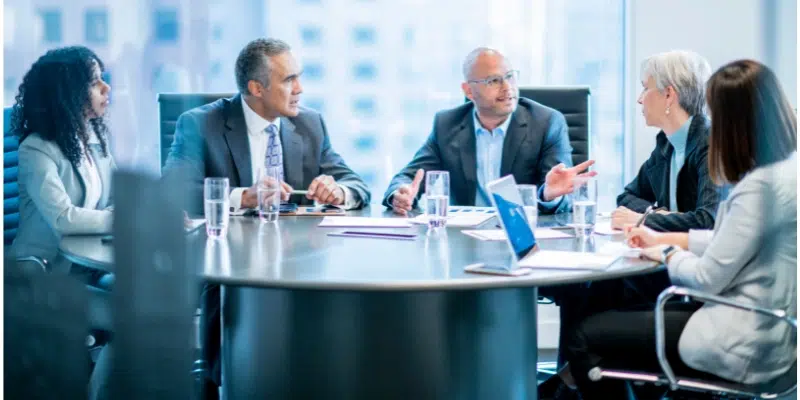 A diverse group of five professionals in a modern conference room, engaged in a meeting around a large circular table.