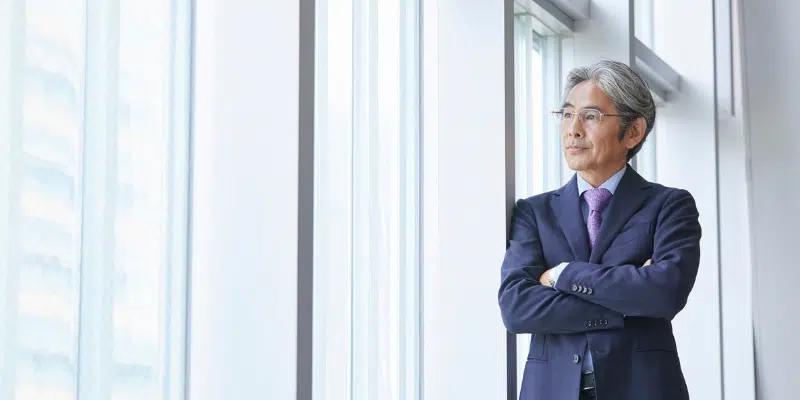An older Asian businessman with gray hair and glasses, standing with arms crossed and looking out a bright window in a modern office