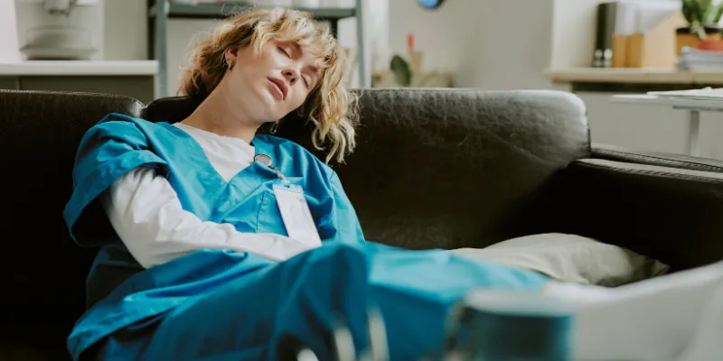 A midwife in blue scrubs asleep on a couch in a hospital staff room