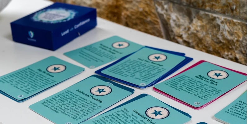 Teal question cards from the Lead with Confidence deck laid out on a white table beside their navy box