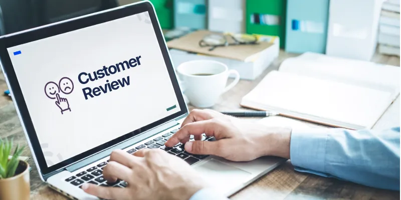 A person typing on a laptop that displays a Customer Review screen with happy and sad face icons, beside a coffee cup and notebook on a desk